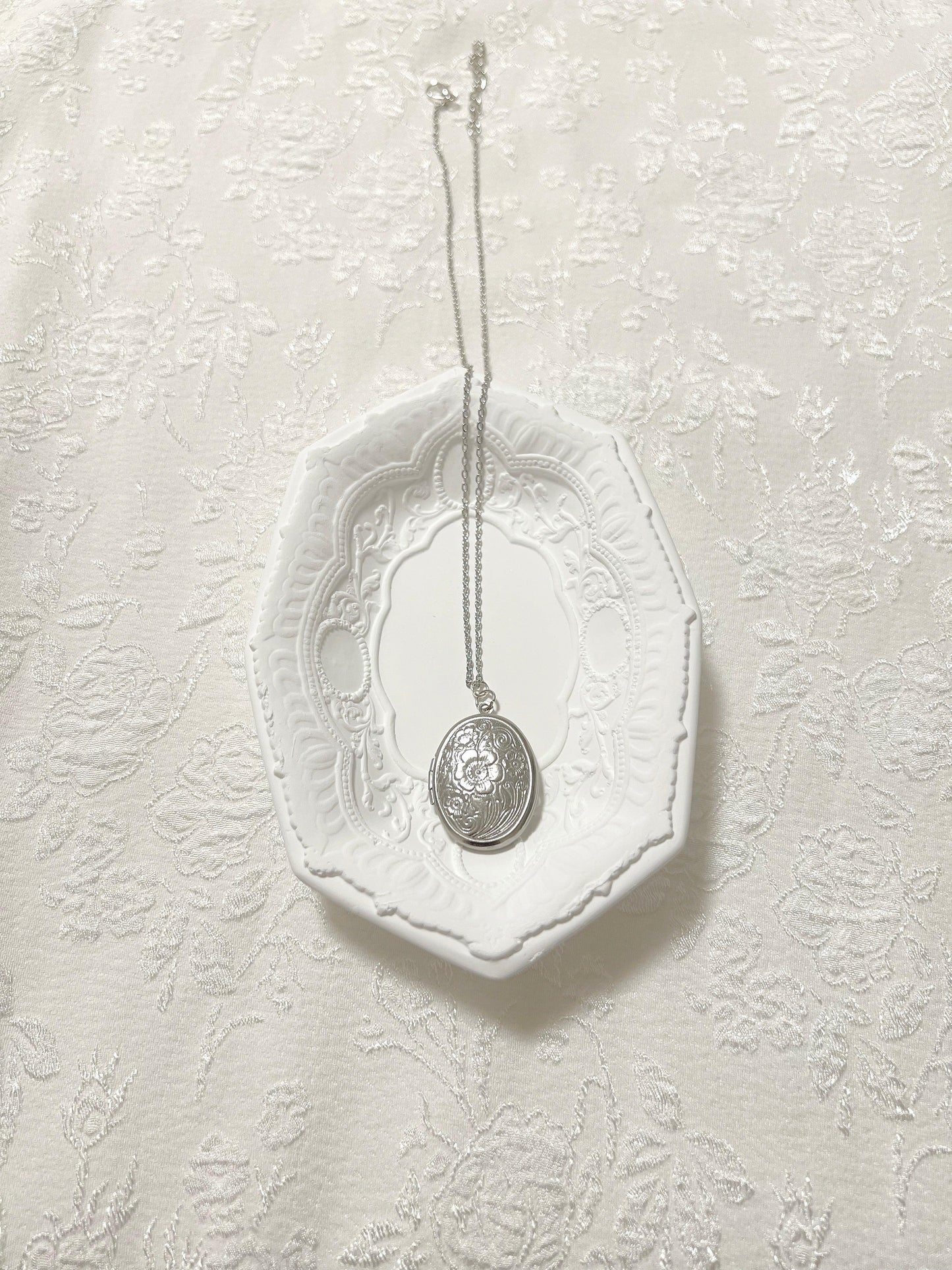 Elegant silver locket necklace with floral engraving, displayed on ornate white dish against textured fabric.
