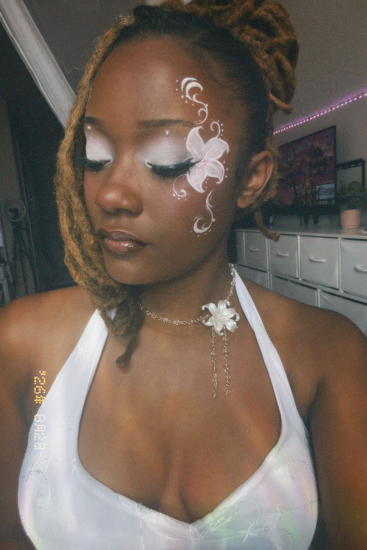 A woman with braided hair and white face paint featuring a butterfly design on her cheek, wearing a white halter top and a white flower necklace in a bedroom setting.