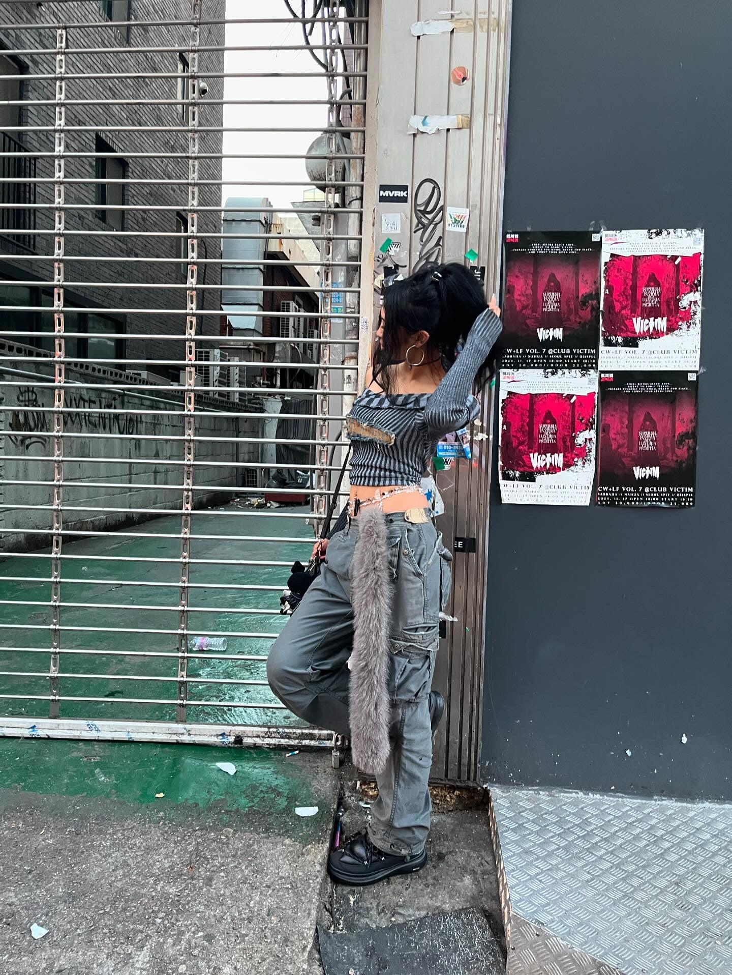 Woman in edgy streetwear with hoop earrings, cargo pants, and faux fur accessory, leaning against urban wall with posters.