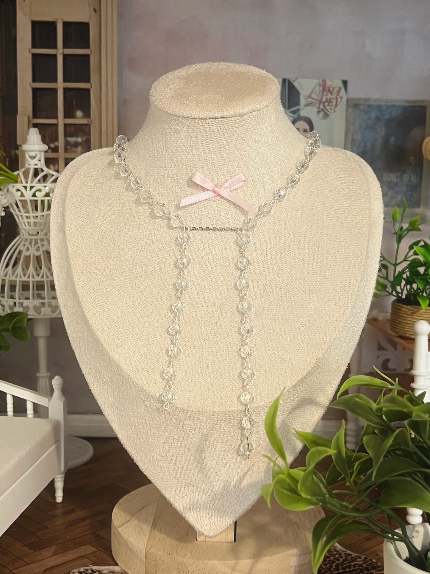 Clear crystal necklace with pink bow, displayed on mannequin with greenery and vintage decor