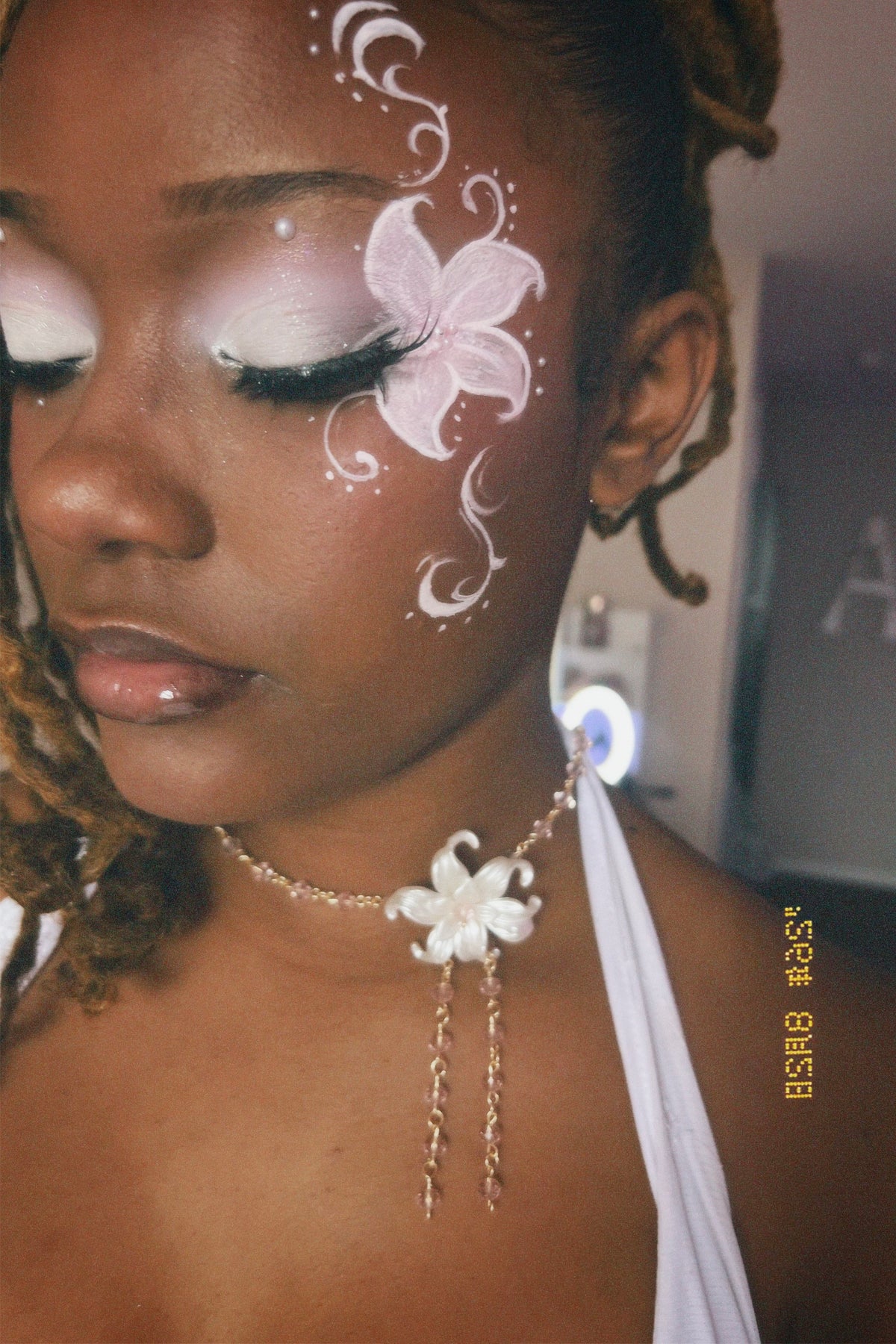 A close-up portrait of a woman wearing a delicate white floral choker necklace with dangling chains. She has elaborate white and pink eye makeup with a butterfly design on her cheek, and her hair is styled in braids.
