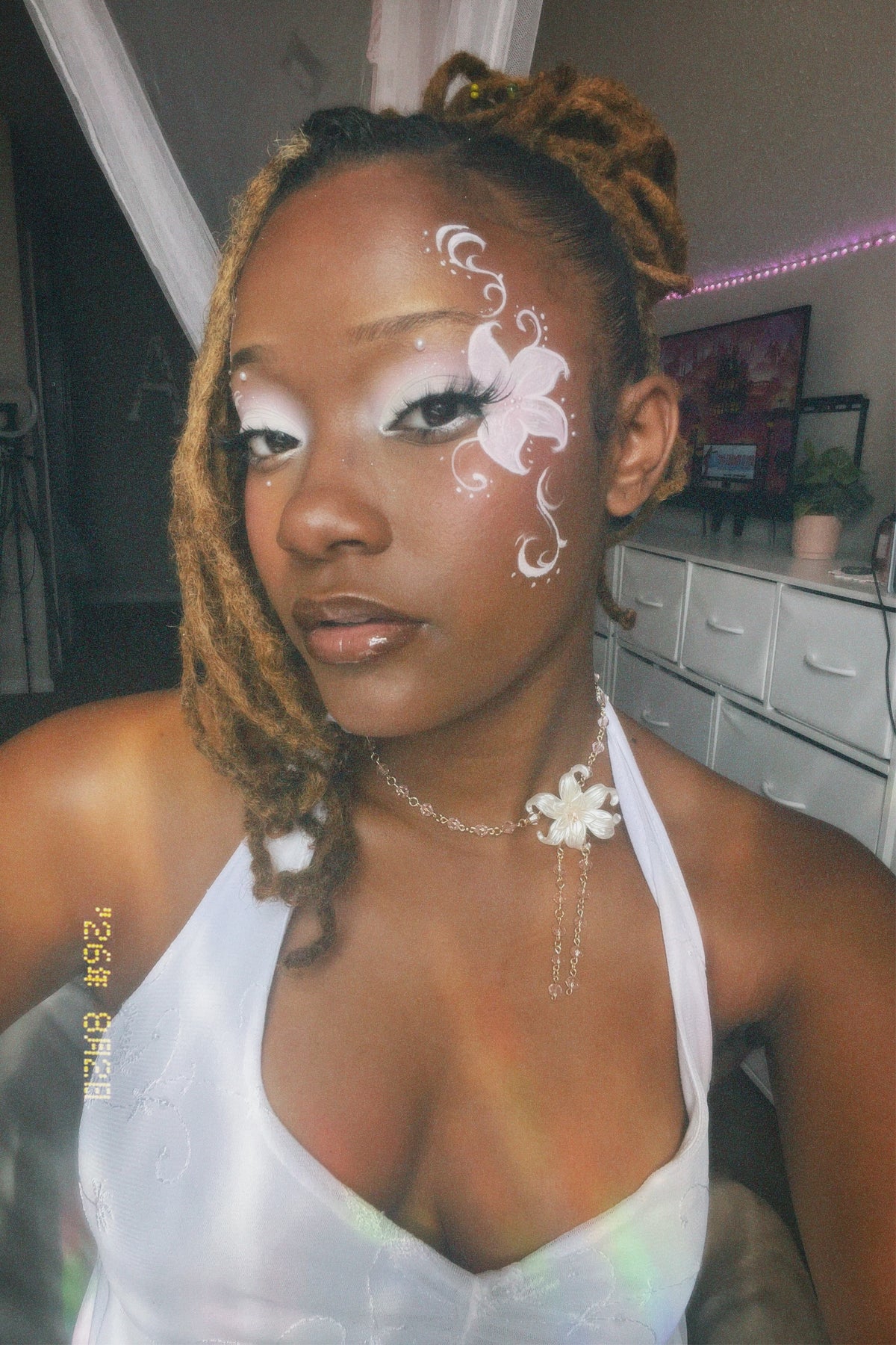 A close-up portrait of a woman with white and pink floral face paint on her cheek, wearing a white halter top and a white flower pendant necklace in a bedroom setting.