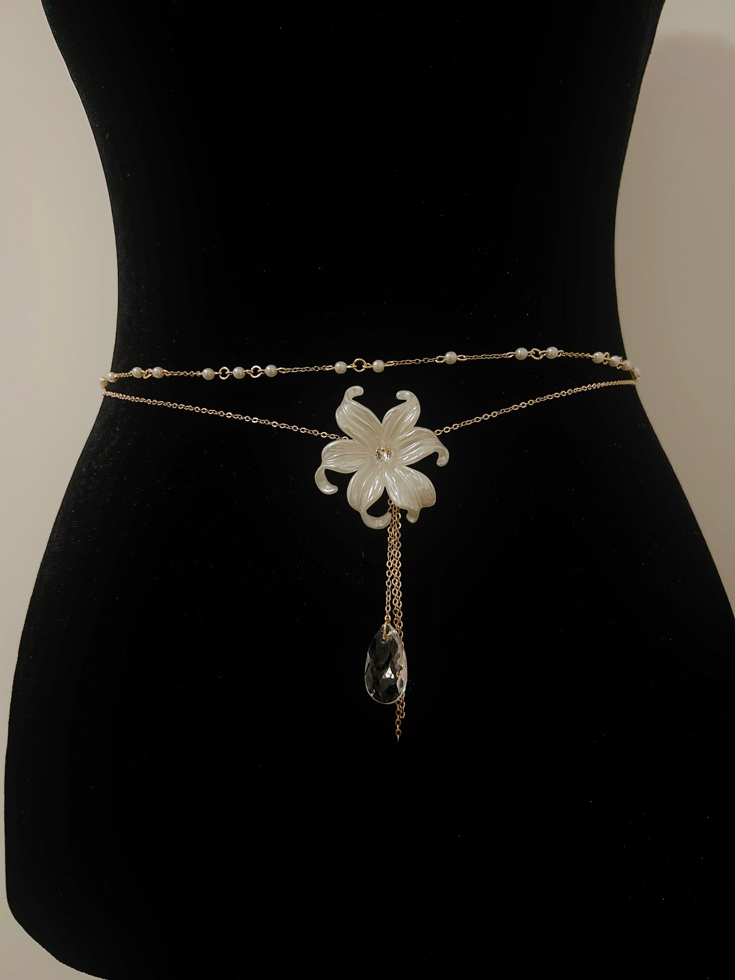 Elegant gold waist chain with white flower pendant, pearl beads, and crystal drop, displayed on black mannequin.