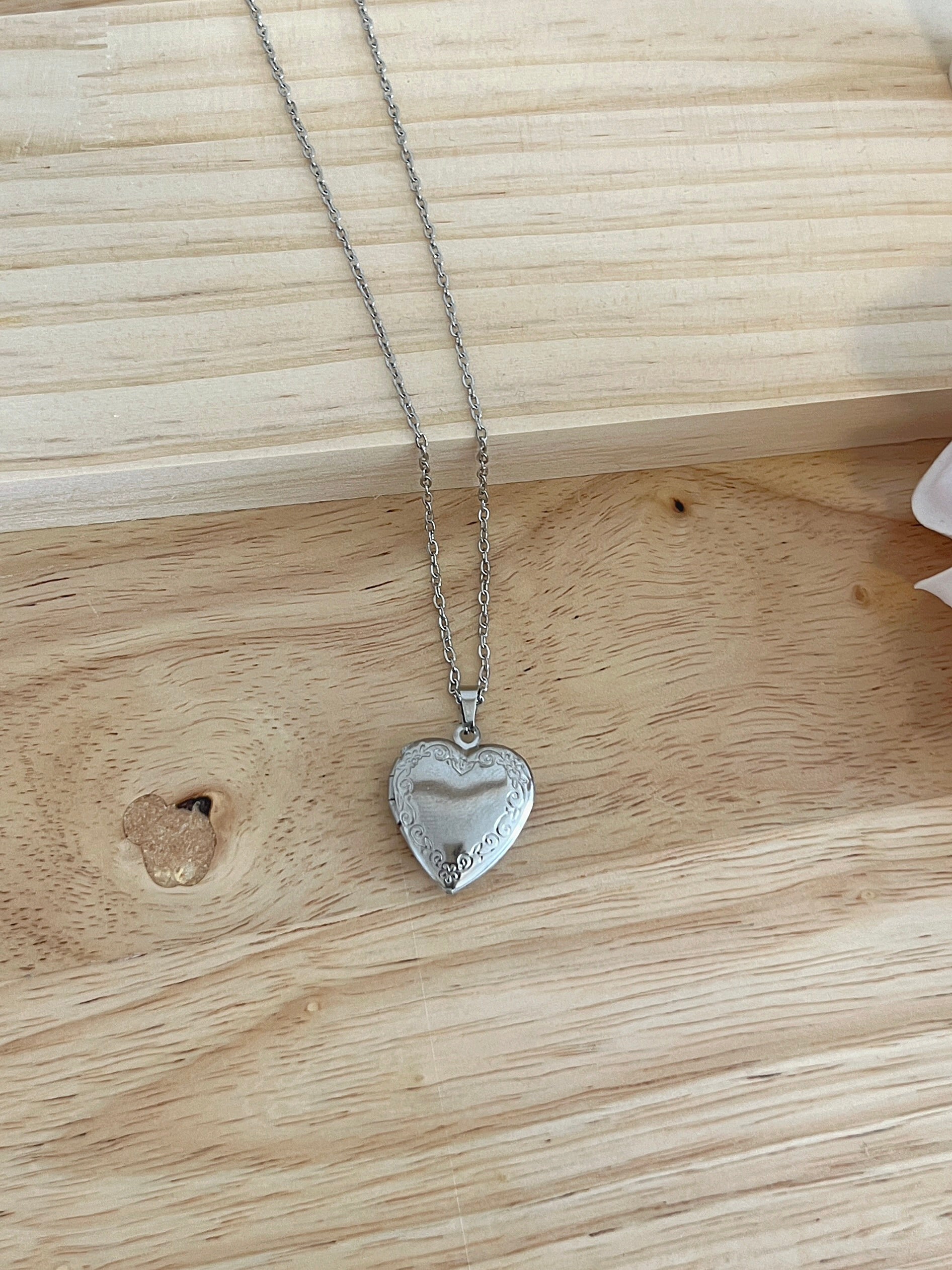 Silver heart locket necklace with intricate scrollwork, displayed on light wood surface.