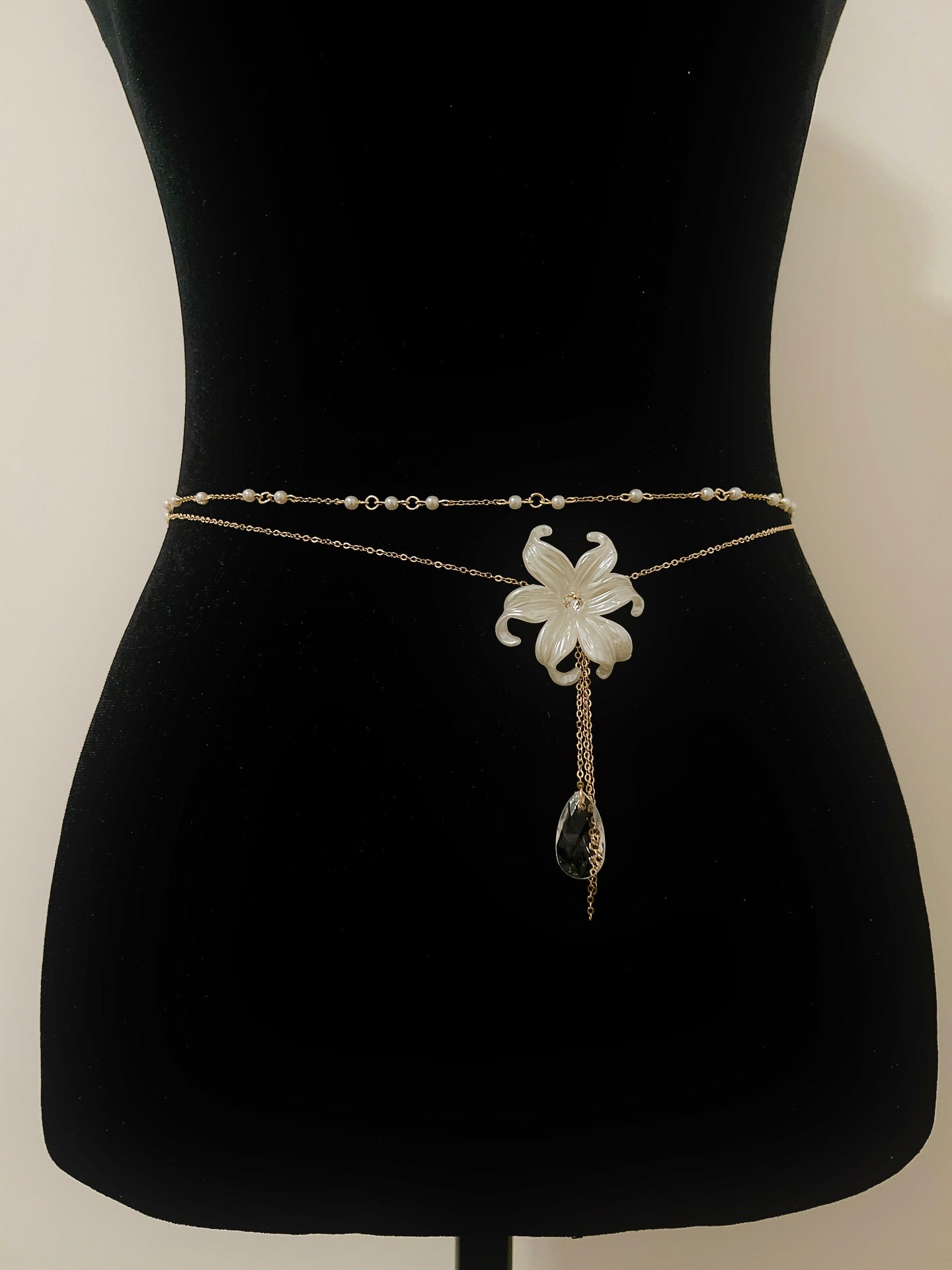 Elegant gold waist chain with white flower pendant, pearl accents, and crystal drop, displayed on black mannequin.