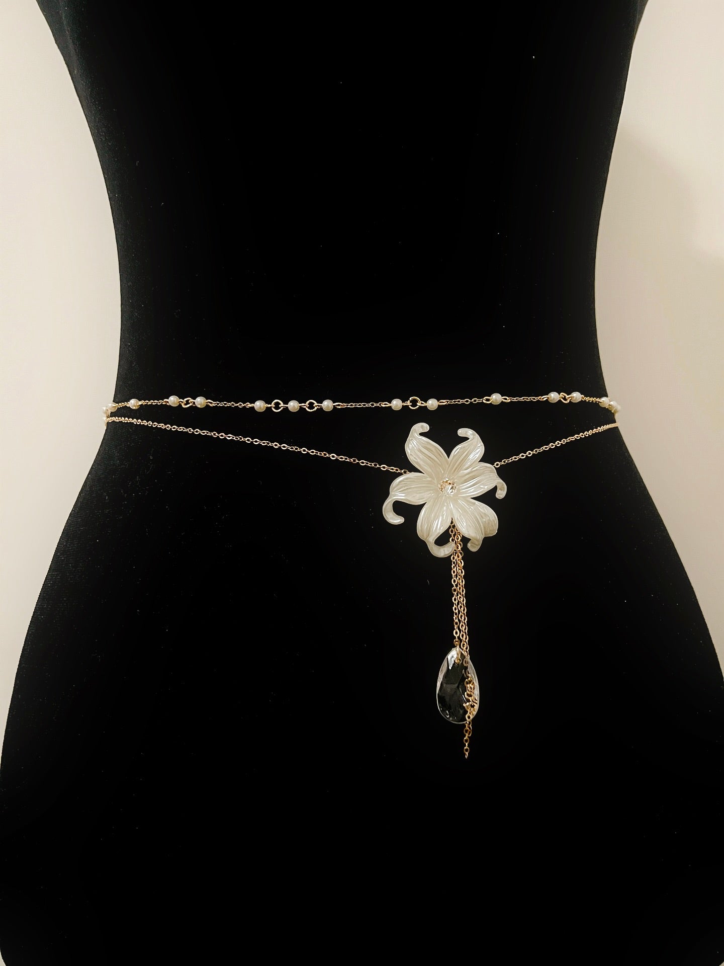 Elegant gold chain waist belt with white flower pendant and crystal drop, displayed on black mannequin.